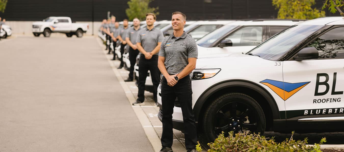 Bluebird Roofing owner standing in front of the company's SUV fleet with his crew lined up behind him